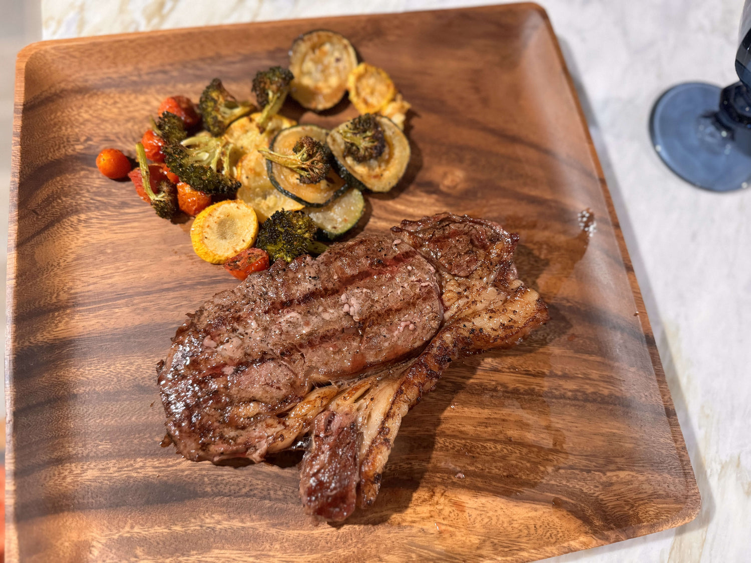 Grilled steak on a wooden plate with roasted broccoli, zucchini, and cherry tomatoes. A glass sits nearby on a marble surface. Warm, inviting tone.