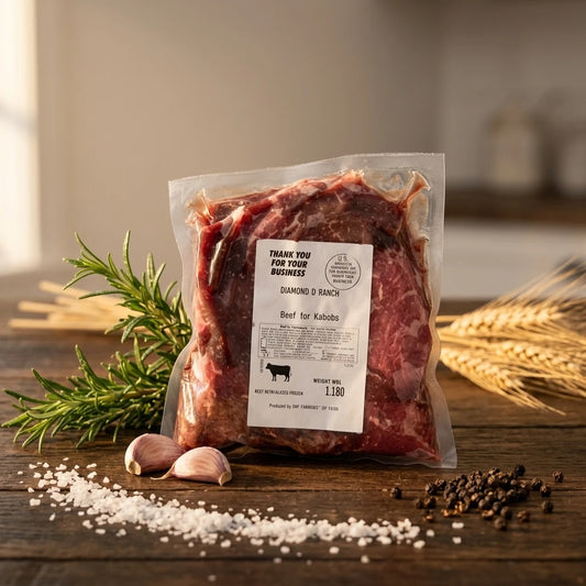 A vacuum-sealed package of raw beef flank steak stands on a rustic wooden surface with salt, peppercorns, garlic, rosemary, and wheat scattered around it.
