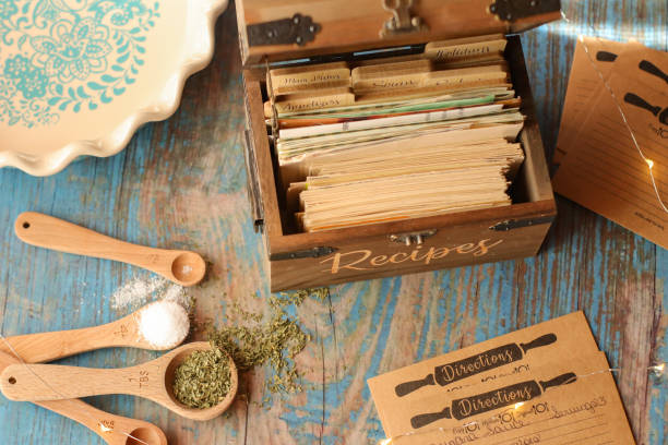 A vintage wooden recipe box filled with organized cards sits on a rustic blue table. Nearby are measuring spoons with herbs and salt, and a decorative ceramic dish.