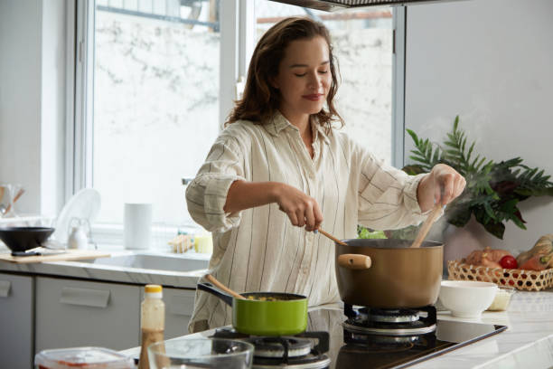 A woman smiles as she cooks in a bright kitchen, stirring a pot on a stove. Fresh ingredients and potted plants add a cozy, inviting vibe.