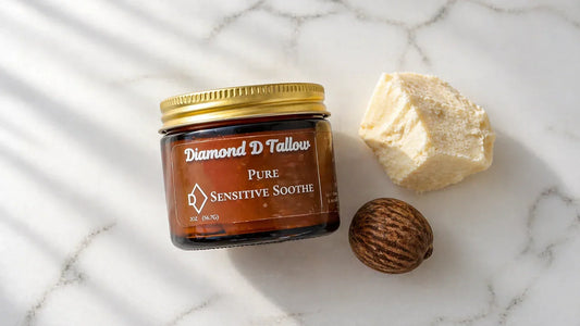 A clean flat-lay on a white marble surface: a small glass jar of creamy white whipped tallow balm sits beside a raw shea nut and a chunk of unrefined shea butter. 