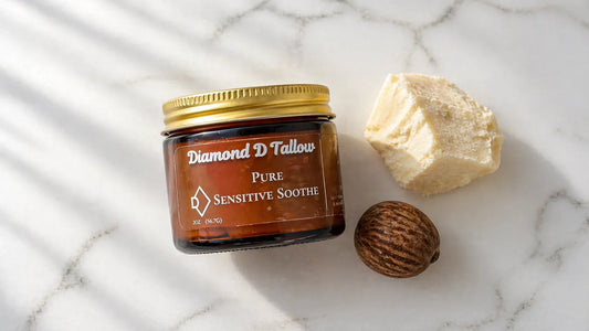 A clean flat-lay on a white marble surface: a small glass jar of creamy white whipped tallow balm sits beside a raw shea nut and a chunk of unrefined shea butter. 