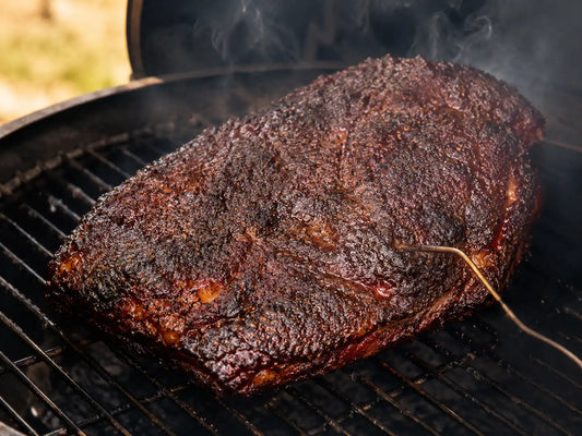 A whole beef brisket resting on a smoker grate, surrounded by thin wisps of white smoke. The bark is dark and fully formed.