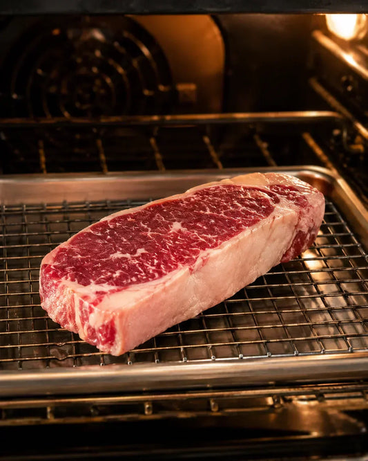 Diamond D Ranch NY strip steak resting on a wire rack over a sheet pan, inside a home oven.