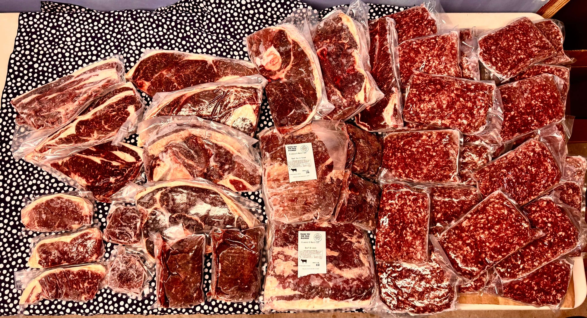 1/8 vacuum-sealed packs of grass-fed beef cuts and ground beef from Diamond D Ranch displayed on a patterned surface.