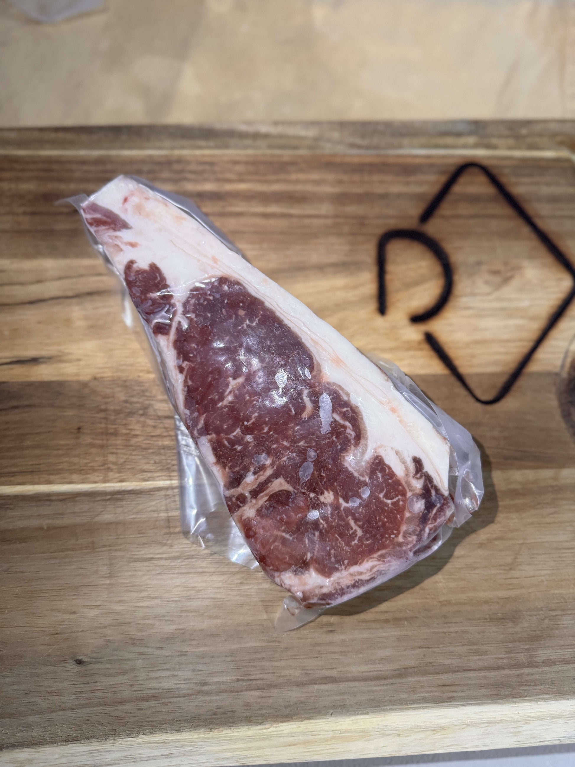A vacuum-sealed ribeye steak on a wooden cutting board. The marbled meat is well-defined against the light wood, creating a fresh and appetizing appearance.