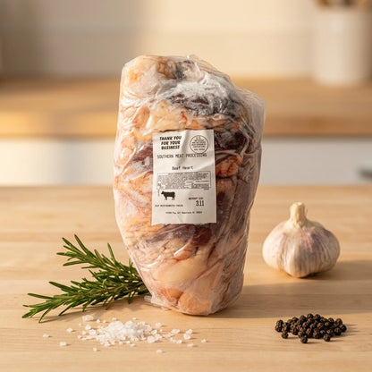 A vacuum-sealed package of grass fed beef heart on a light wood surface, with a garlic bulb, rosemary sprig, coarse salt, and black peppercorns