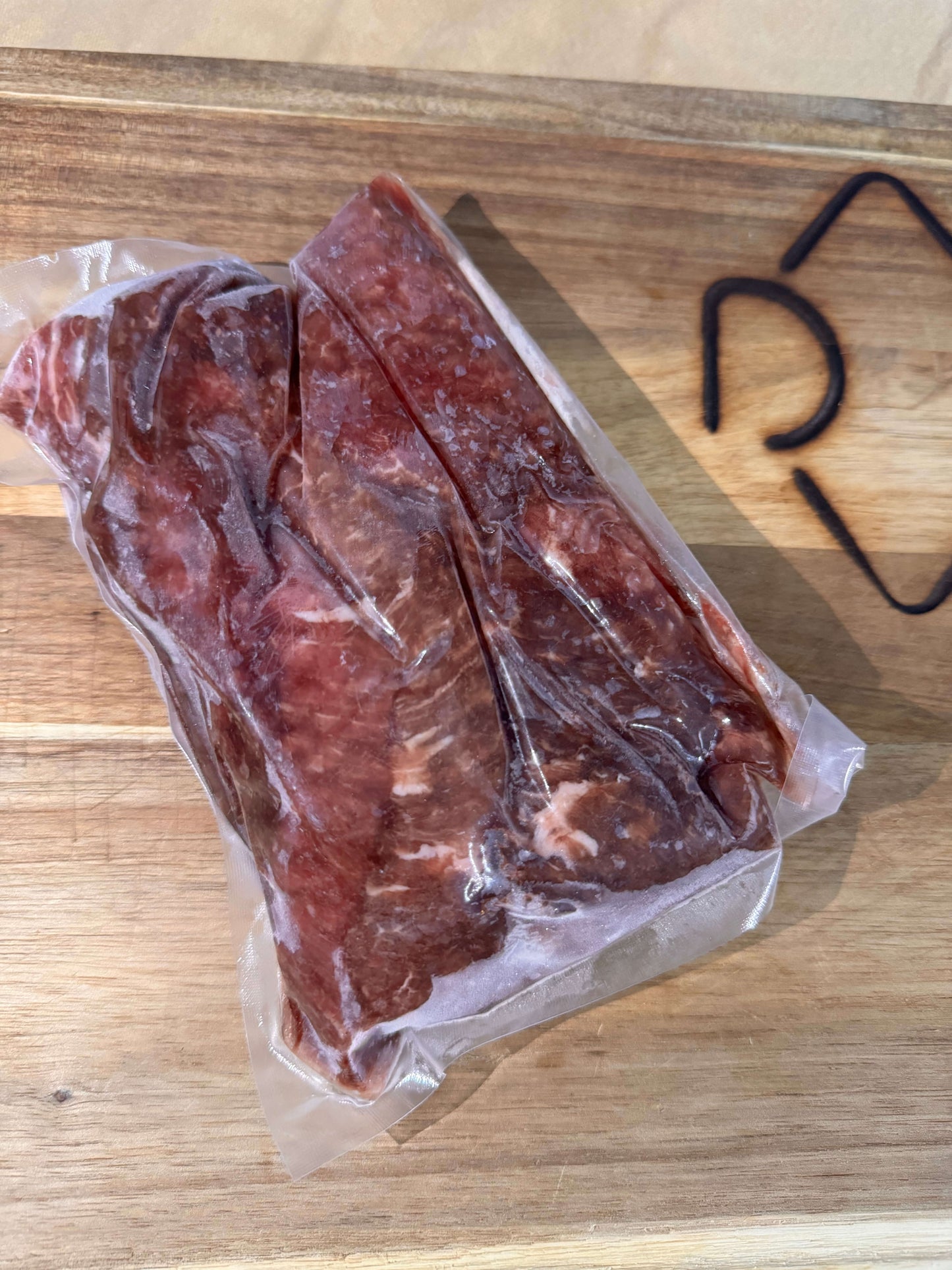 Frozen cubed steak in vacuum-sealed plastic on a wooden cutting board, next to a partially visible black design, indicating freshness and quality.