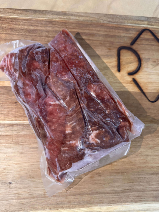 Frozen cubed steak in vacuum-sealed plastic on a wooden cutting board, next to a partially visible black design, indicating freshness and quality.