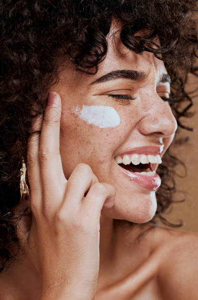 A woman with curly hair smiles broadly, applying whipped tallow to her cheek with her finger. Her expression is joyful and the setting is warm and relaxed.