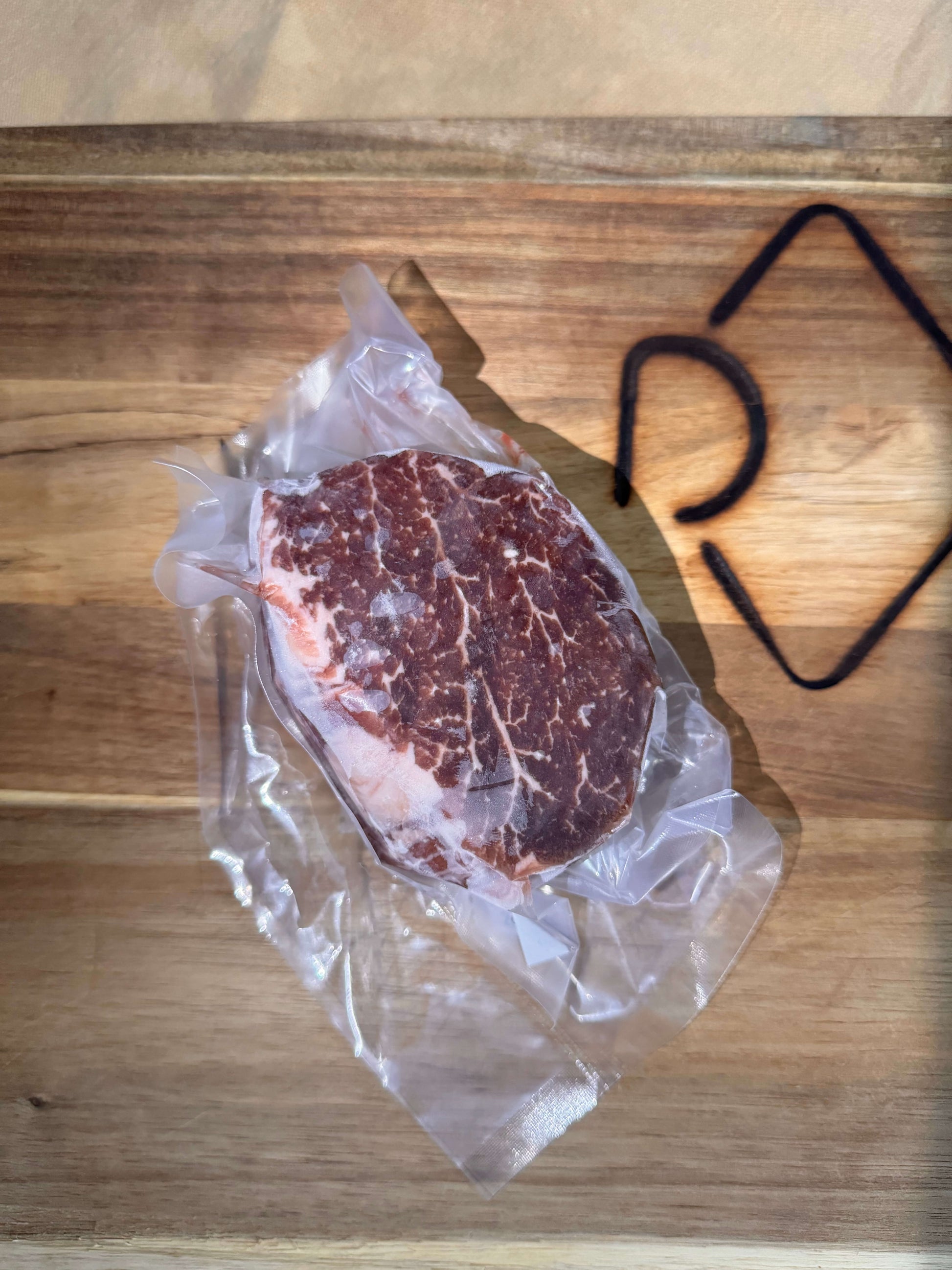 Vacuum-sealed marbled filet mignon steak on a wooden cutting board; the sun creates a geometric shadow. The raw meat is rich in color, conveying freshness.