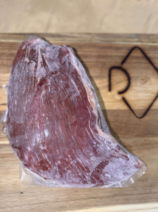 A frozen, vacuum-sealed flank steak rests on a wooden cutting board with a geometric scribe in the corner. The steak appears raw and marbled.