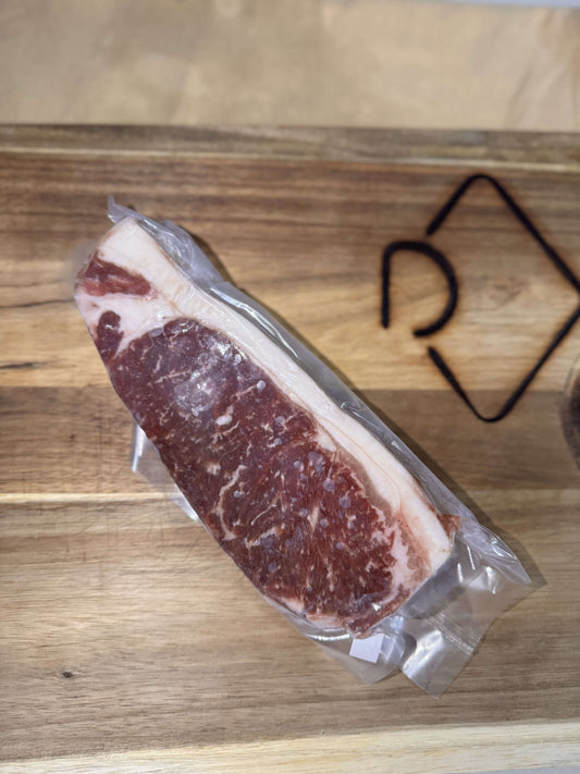 A vacuum-sealed raw NY Strip steak with marbled fat lies on a wooden cutting board. The setting is simple, highlighting the fresh quality of the meat.