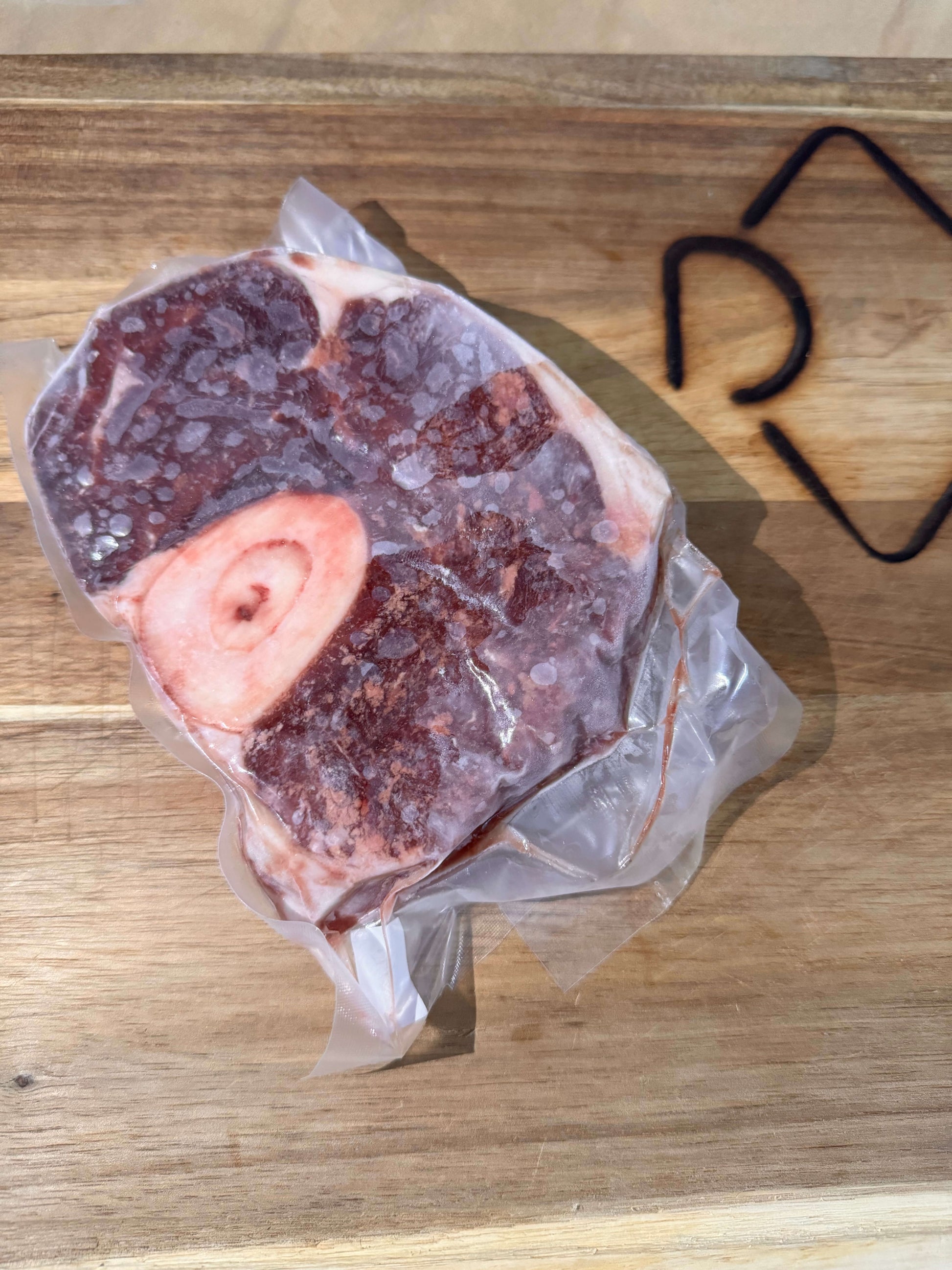 A vacuum-sealed package of frozen osso buco meat with a bone is placed on a wooden cutting board. The meat is frosty, indicating it's very cold.