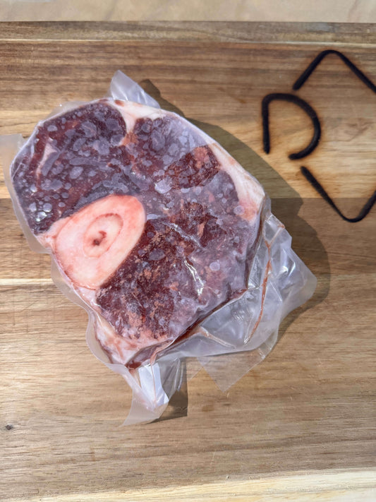 A vacuum-sealed package of frozen osso buco meat with a bone is placed on a wooden cutting board. The meat is frosty, indicating it's very cold.