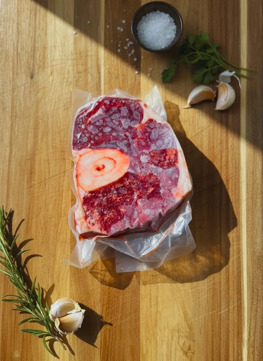 A vacuum-sealed package of frozen osso buco meat with a bone is placed on a wooden cutting board. The meat is frosty, indicating it's very cold.