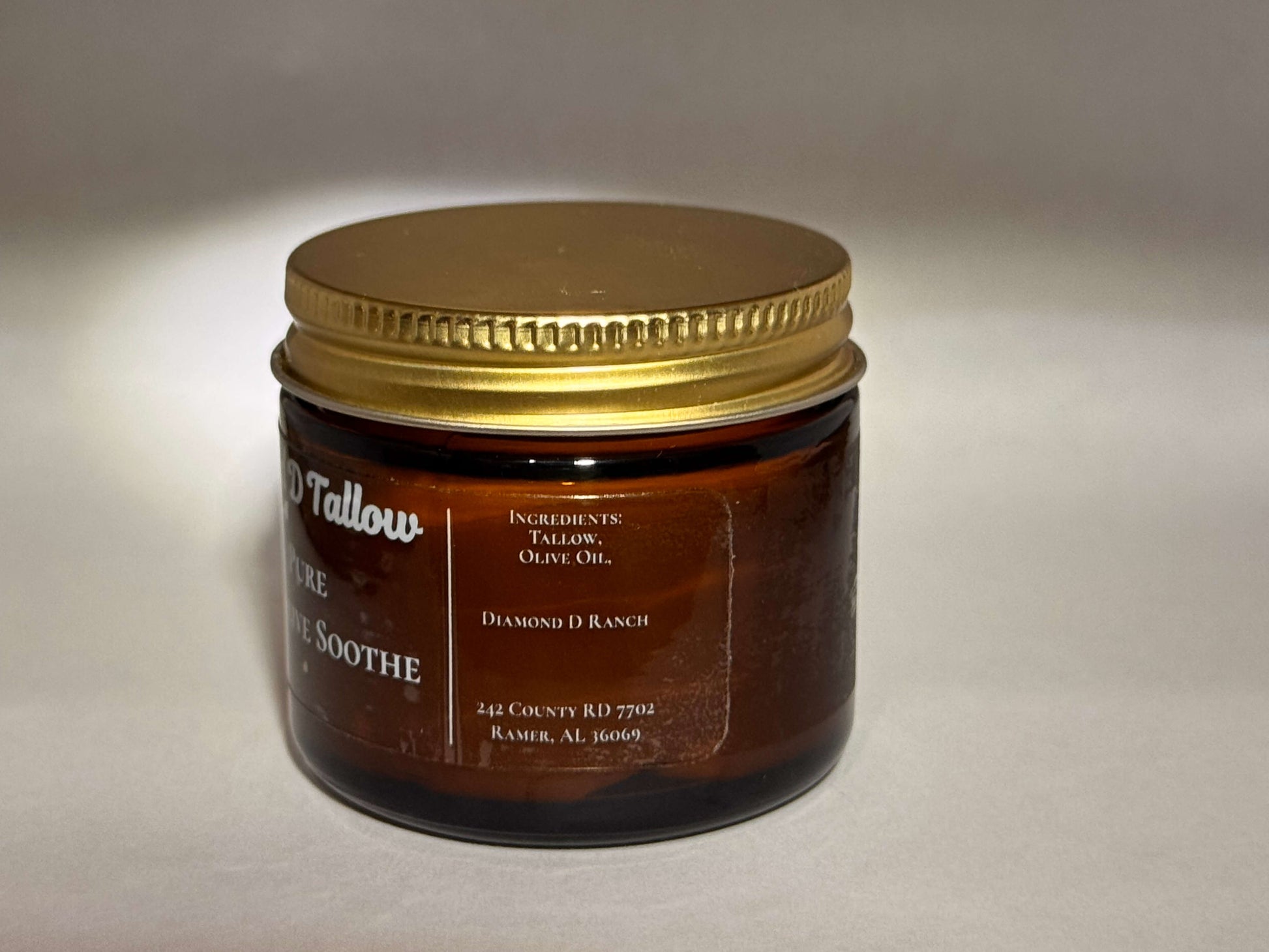 A brown glass jar with a gold lid, labeled as "Diamond D Tallow." Contains tallow and olive oil. Text shows location in Ramer, AL. Simple and elegant.