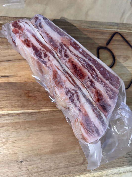 A vacuum-sealed slab of marbled short ribs placed on a wooden cutting board. The short ribs have layers of fat and meat which are visible through the plastic wrapping.