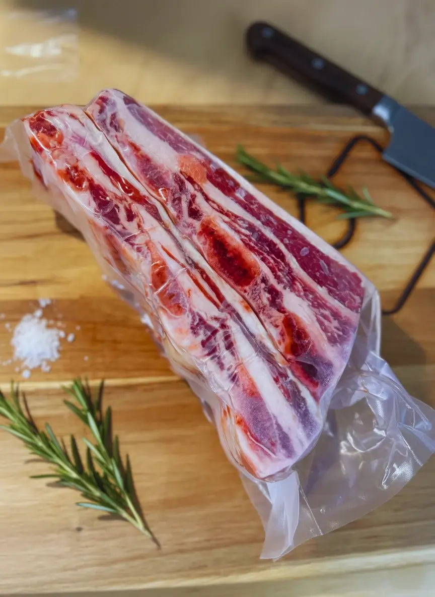 A vacuum-sealed slab of marbled short ribs placed on a wooden cutting board. The short ribs have layers of fat and meat which are visible through the plastic wrapping.