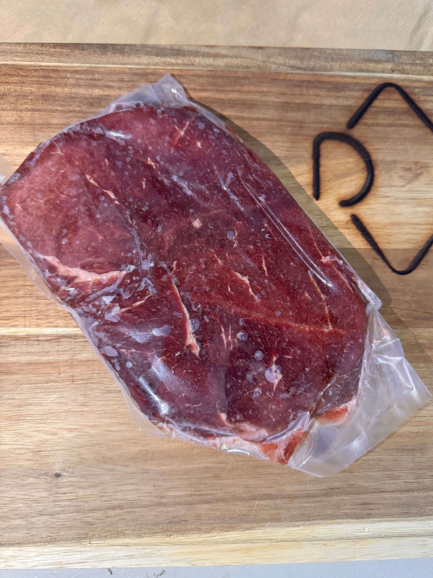 A raw sirloin steak sealed in plastic wrap on a wooden cutting board, bathed in natural light. The texture of the meat and clear packaging are visible.