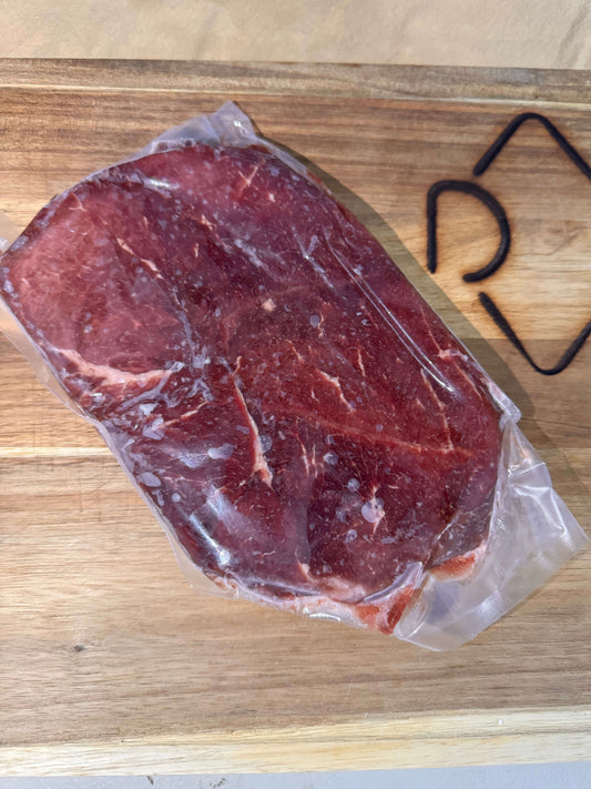 A raw sirloin steak sealed in plastic wrap on a wooden cutting board, bathed in natural light. The texture of the meat and clear packaging are visible.