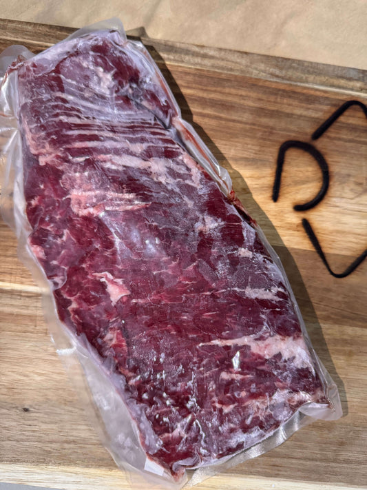 A slab of raw Skirt Steak in vacuum-sealed plastic is on a wooden cutting board. The marbling is visible, and a faint steak branding is seen beside it.