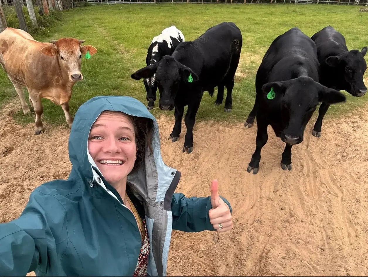 Sydney is in a raincoat, giving a thumbs-up with cows in the background at Diamond D Ranch