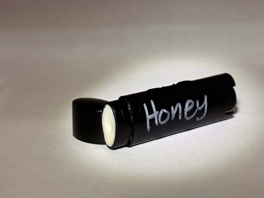 A black cylindrical container for tallow chapstick with "Honey" written in white text is centered against a soft, neutral background with a side-view angle. The lighting is gentle and muted.