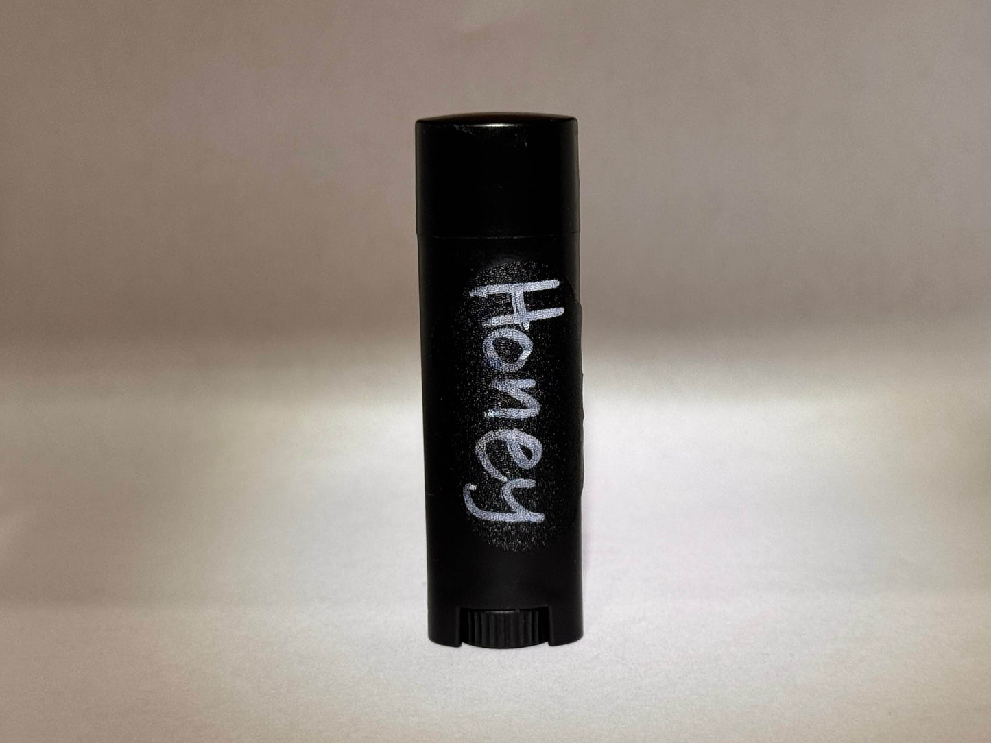 A black cylindrical container for tallow chapstick with "Honey" written in white text is centered against a soft, neutral background. The lighting is gentle and muted.