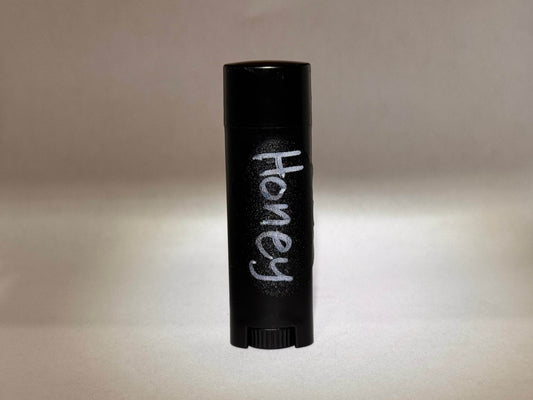 A black cylindrical container for tallow chapstick with "Honey" written in white text is centered against a soft, neutral background. The lighting is gentle and muted.