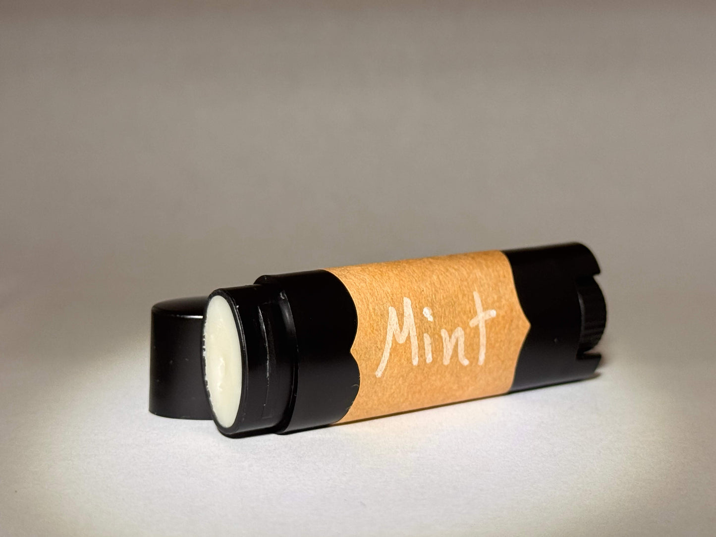 A black tube of lip balm lies on its side with a clear cap, featuring an orange label that reads "Mint." The background is softly lit, creating a cozy, inviting feel.