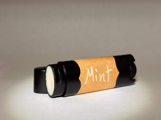 A black tube of lip balm lies on its side with a clear cap, featuring an orange label that reads "Mint." The background is softly lit, creating a cozy, inviting feel.