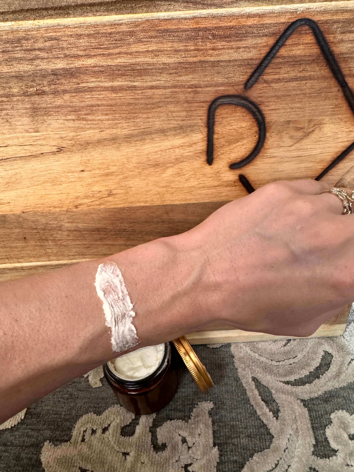 A hand applying white cream onto the forearm over a wooden surface with decorative black metal accents. An open jar with a whipped tallow rests nearby.